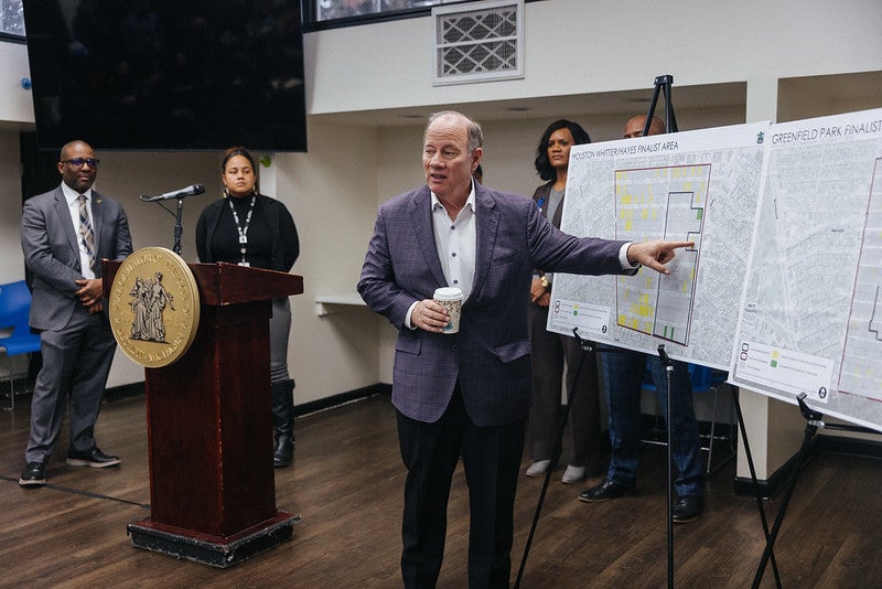 A white man in a gray suit pointing out details on a posterboard. In the background are four more individuals.