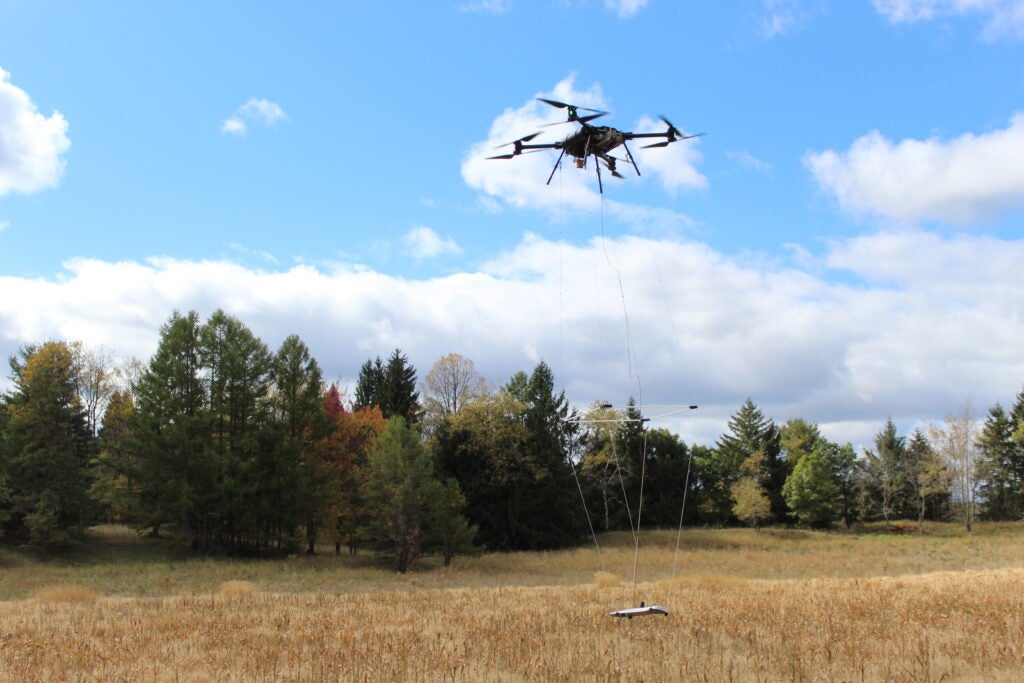 Drone carrying a magnetometer takes flight over a field in Pennsylvania. Autumn treeline is visible in the background against a blue sky and clouds.