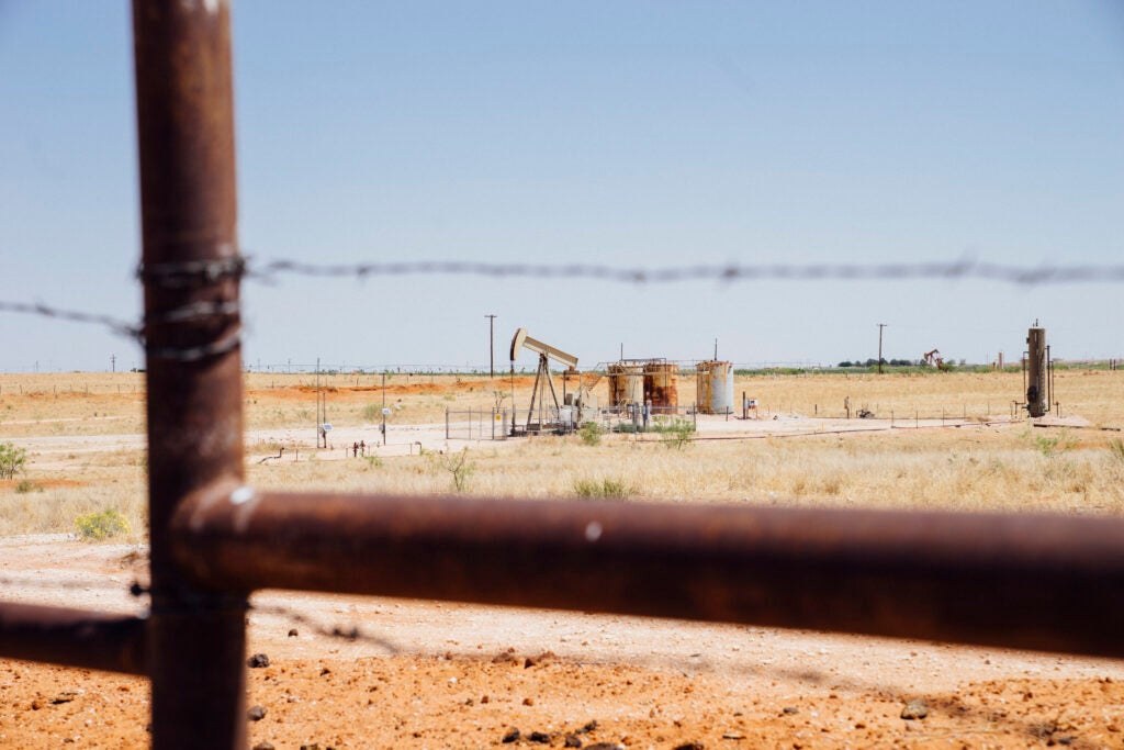 View of a pumpjack at a wellsite in Hobbs, NM. Foreground is a metal fence with barbed wire.