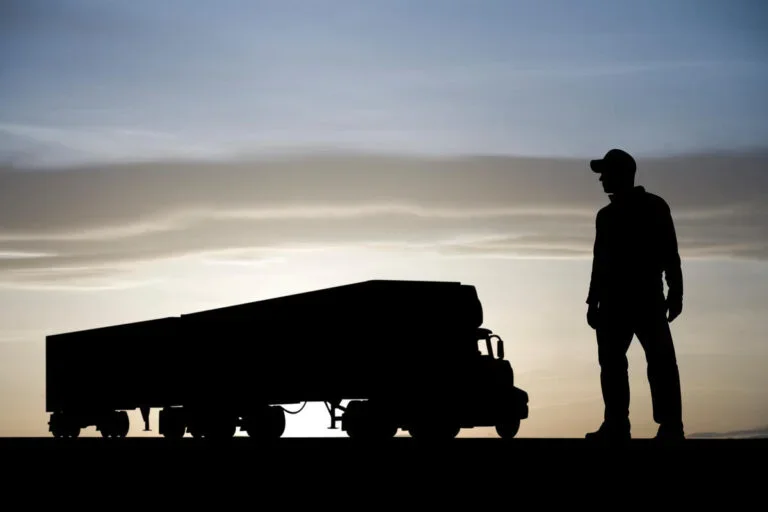 truck driver standing in the foreground with his tractor trailer rig in the background. The image is against a setting sun.
