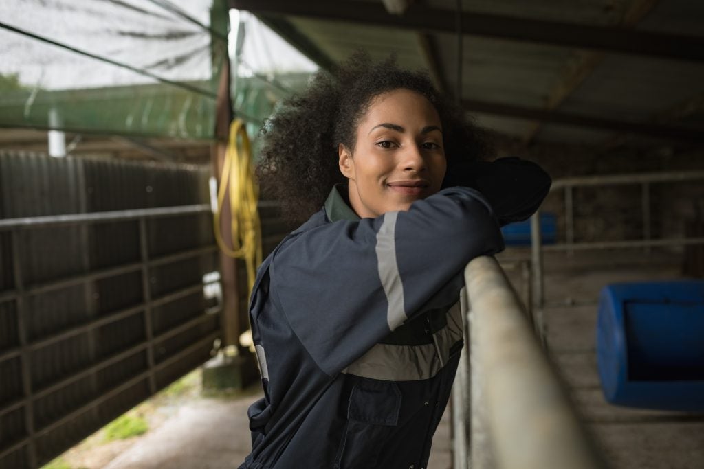 A Black female farmer and/or rancher looks at the camera while leaning on the railing around an animal pen. Her hair is in a high ponytail, and she is wearing a blue jumpsuit.