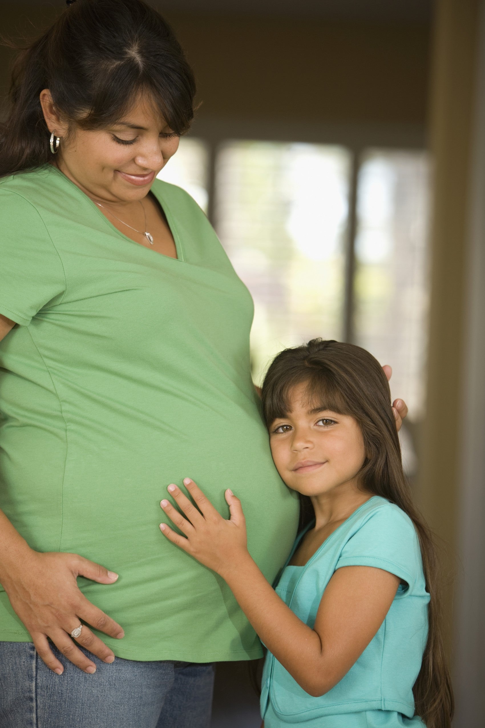 Pregnant Latine woman gazing lovingly at young daughter who is hugging her belly.