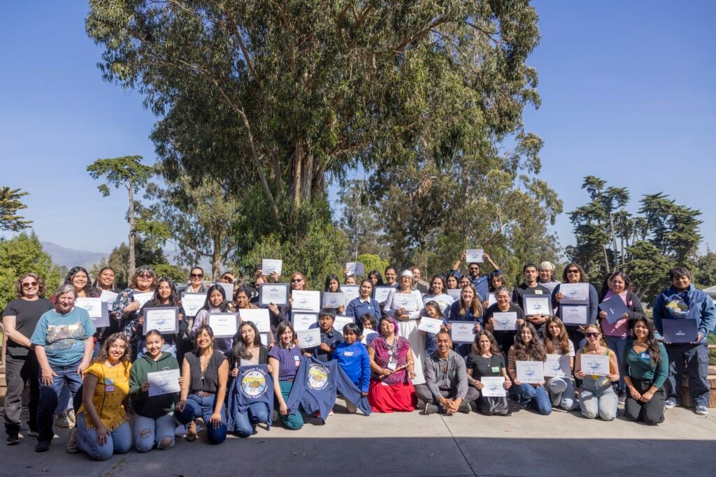 Roughly 50 WLI participants hold their certificates of completion beneath a tree outside.